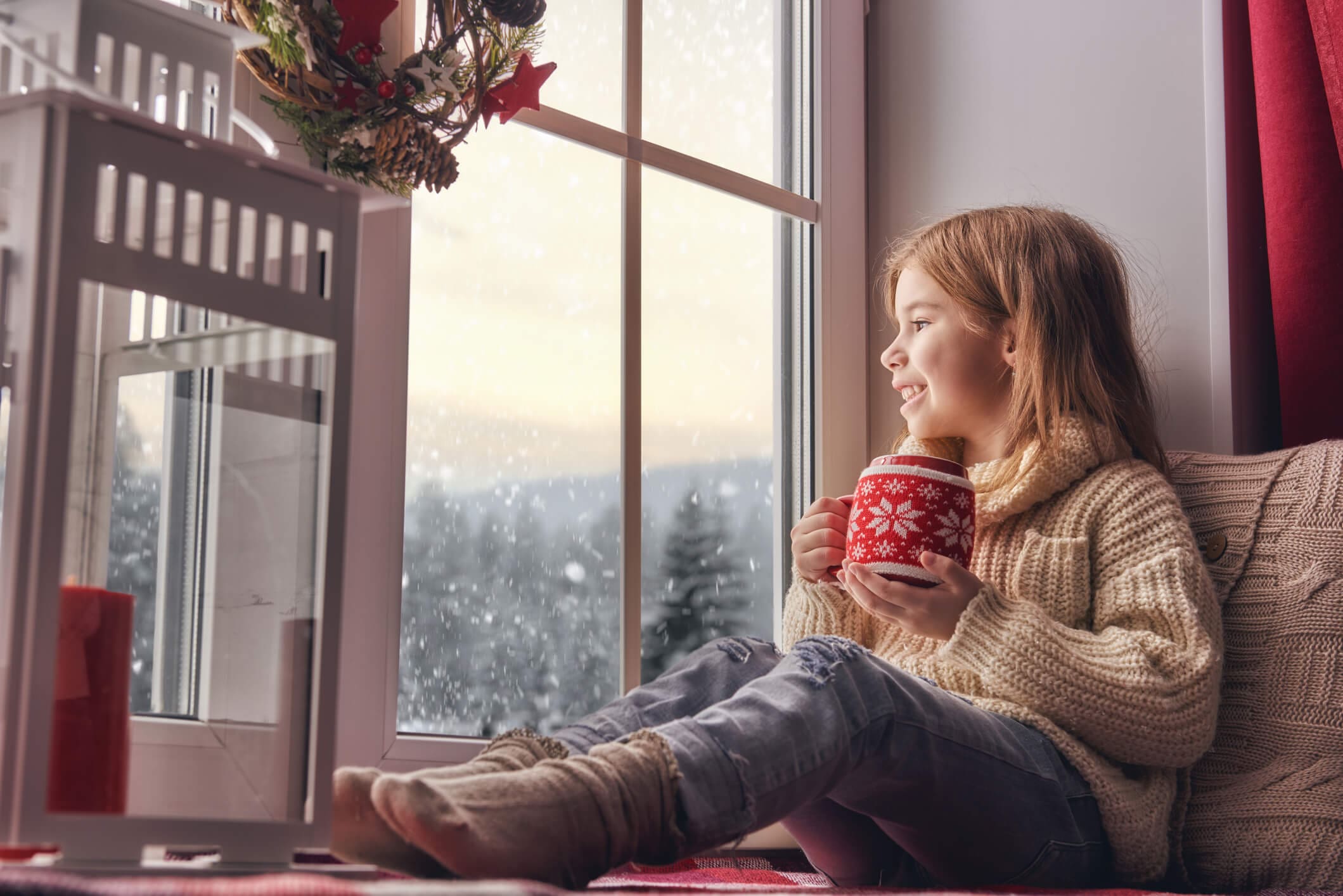 Girl sitting by window. Merry Christmas and happy holidays! Cute little girl sitting by the window with a cup of hot drink and looking at the winter forest. Room decorated on Christmas. Kid enjoys the snowfall.